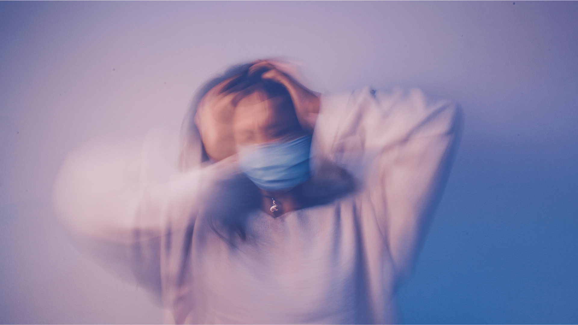 A blurred figure wearing a blue face mask holds their head with both hands against a soft purple background, conveying stress or anxiety.